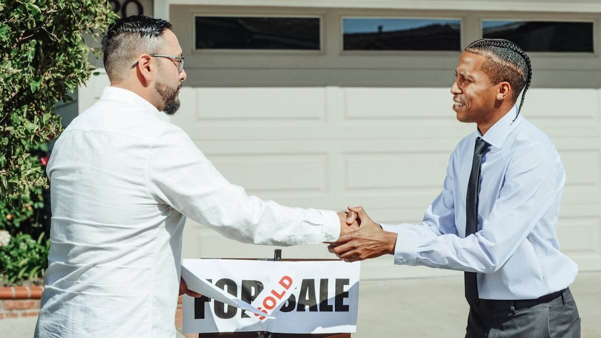 Two men shaking hands in front of a sold house sign after a successful home sale